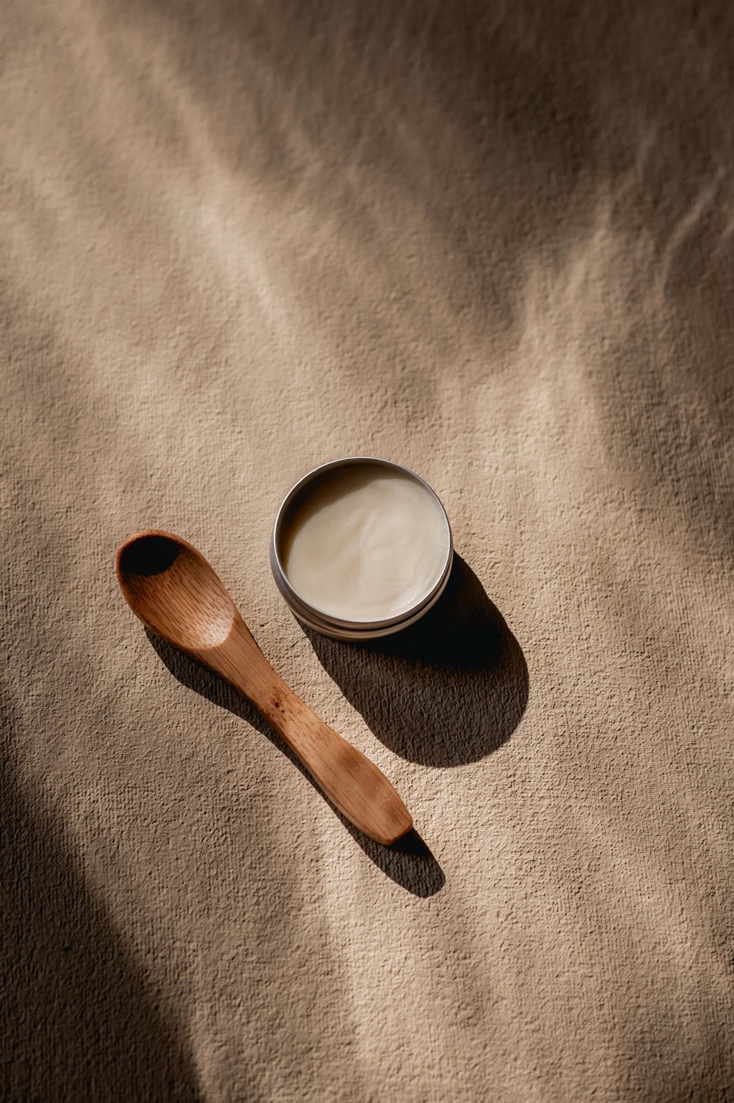 Houten cosmetica lepeltje van 10 cm naast een blik shea boter op een zachte ondergrond.