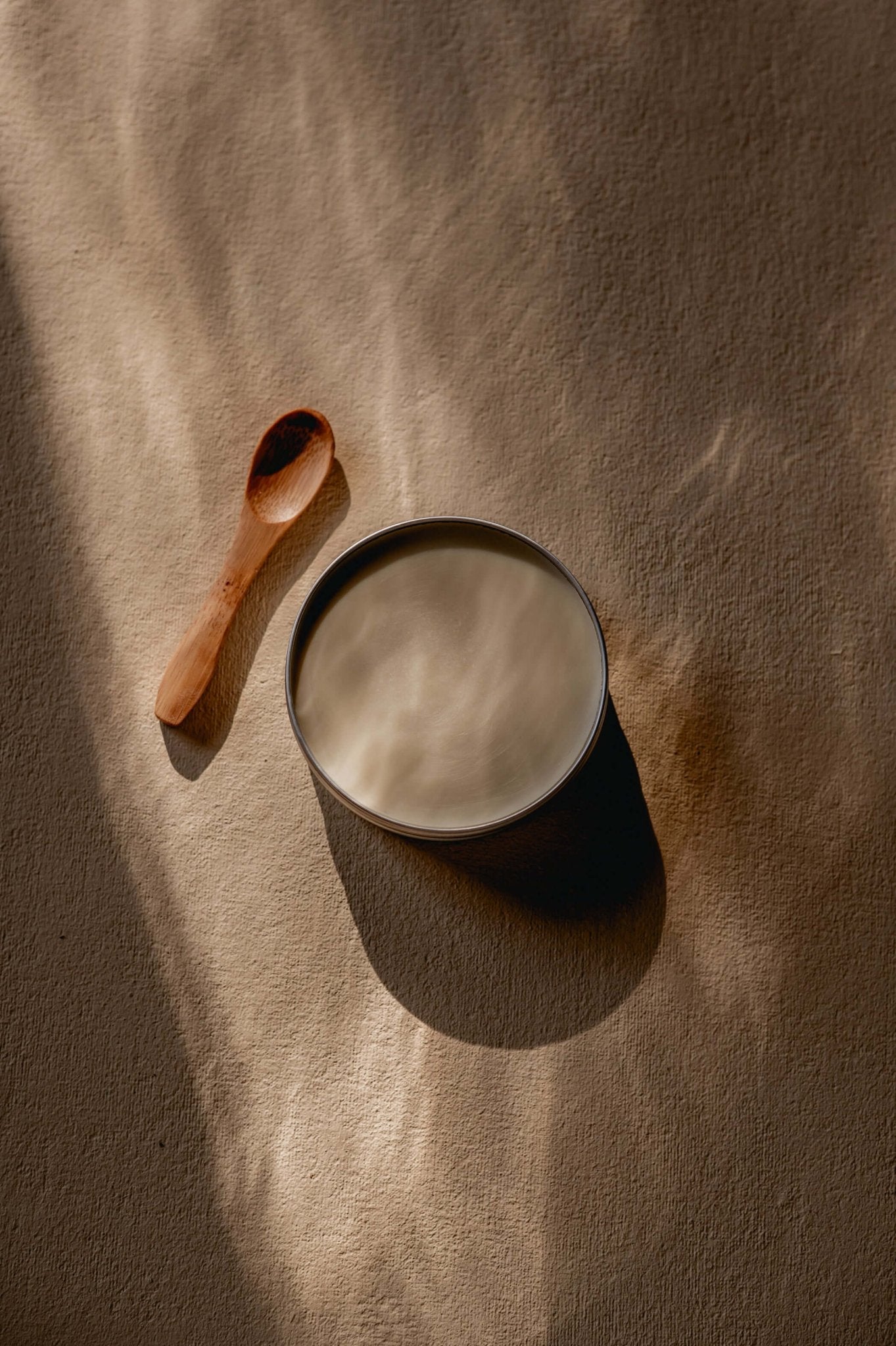 Namashea open blik shea boter met houten cosmetica lepeltje, close-up vanuit een andere hoek op een zachte ondergrond