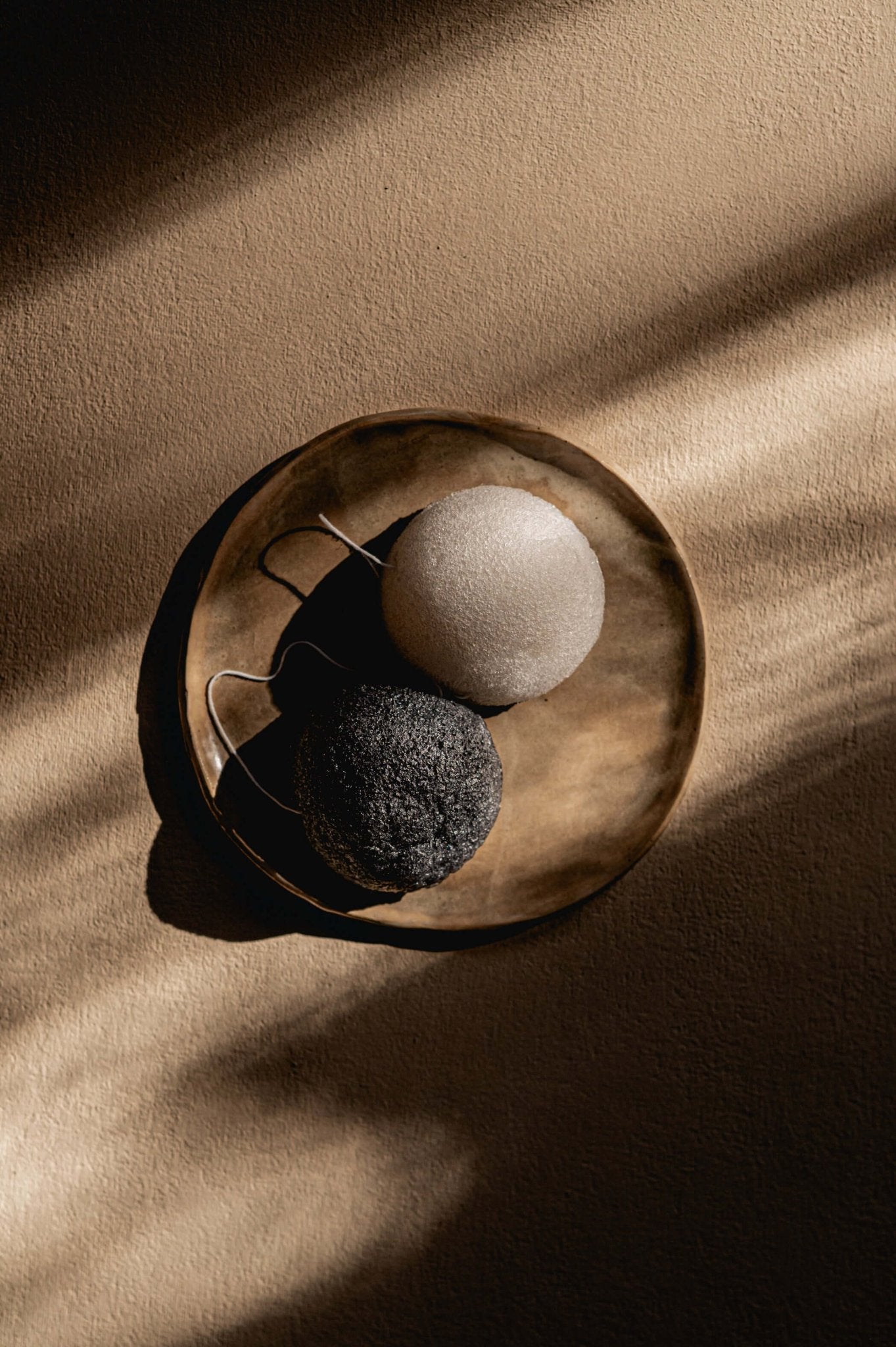 Namashea konjac sponzen Puur (wit) en Houtskool (zwart) naast elkaar op een schaaltje, close-up op een beige ondergrond.