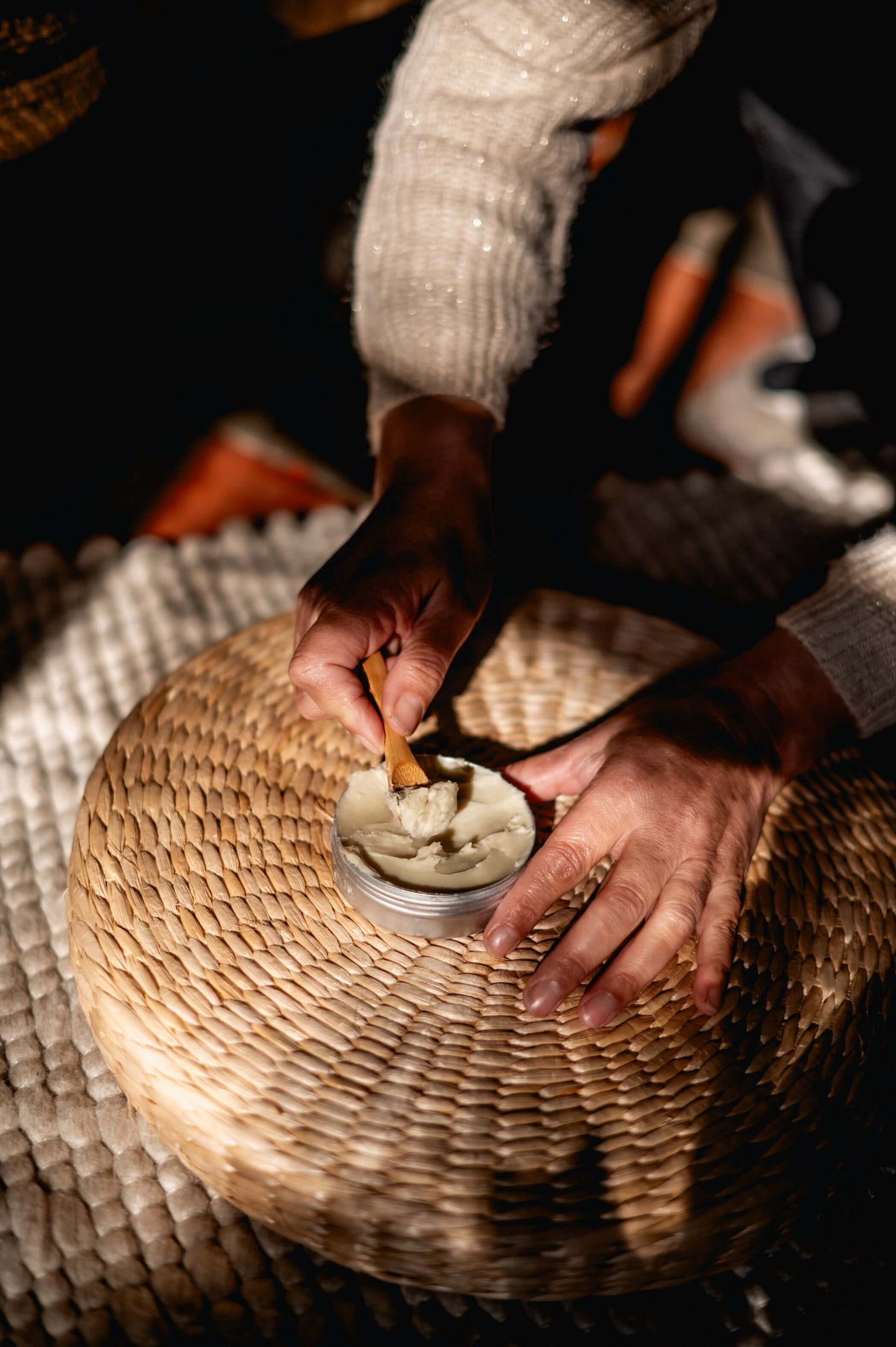 Applying shea butter from a tin with a wooden stick on a woven surface, showcasing natural skincare use.