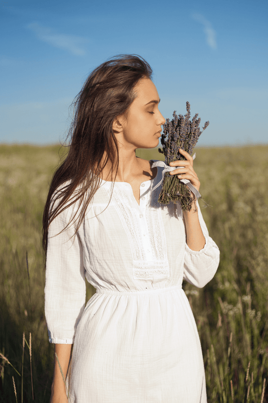 Vrouw in witte jurk ruikt aan een bosje lavendel in een natuurlijk veld bij daglicht