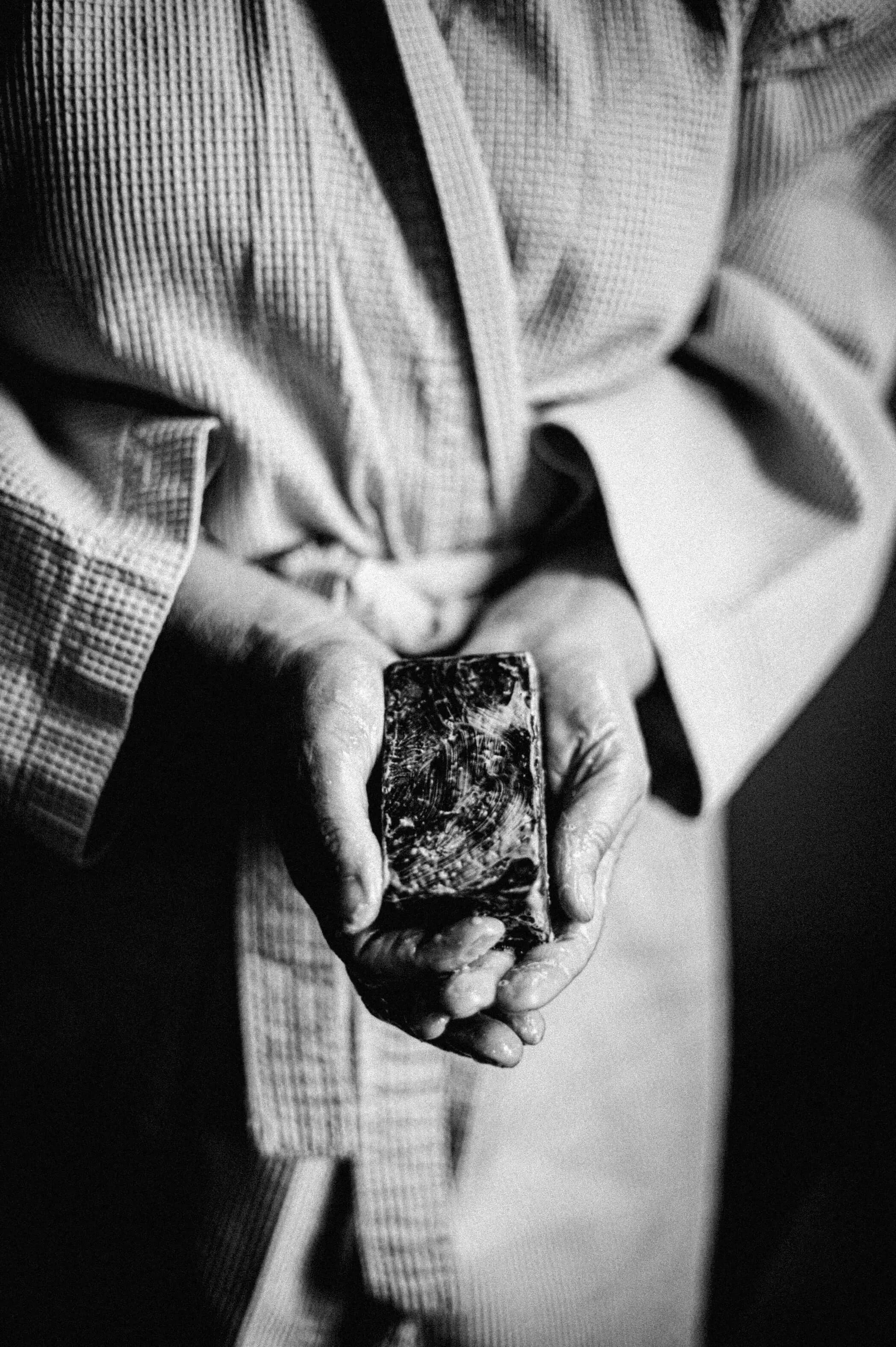 Zwart-wit sfeerbeeld van een persoon in badjas die een schuimend blok Namashea African Black Soap vasthoudt.