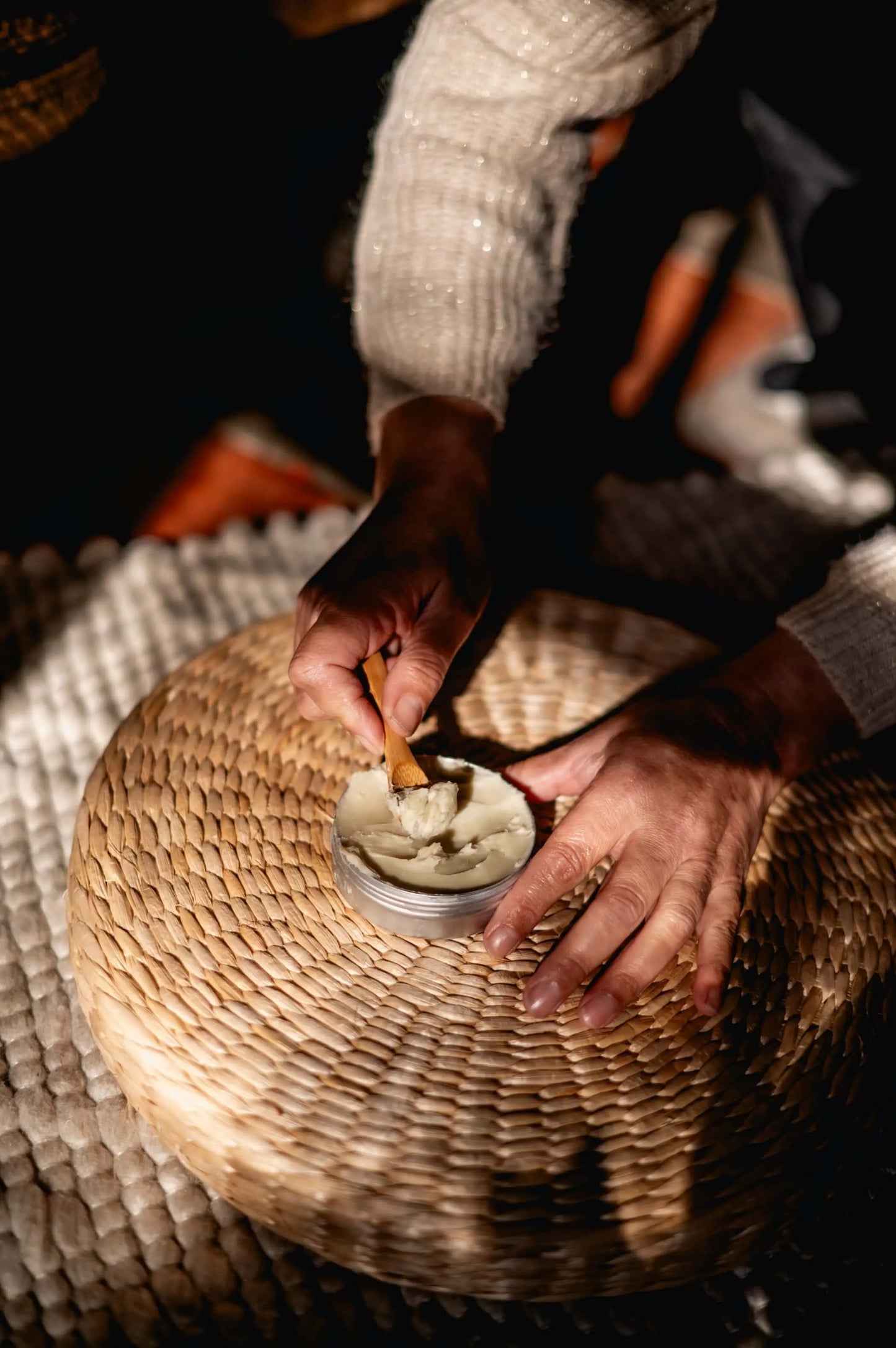 Applying shea butter from a tin with a wooden stick on a woven surface, showcasing natural skincare use.