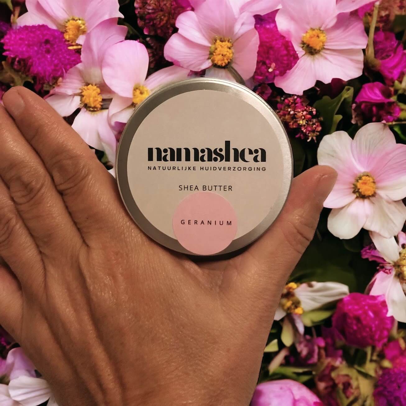 blik shea boter van Namashea tussen duim en wijsvinger. shea boter met geraniumolie met achtergrond vol rose bloemen